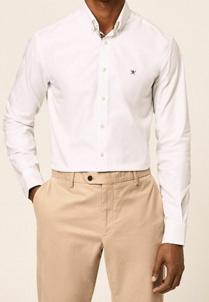 Man draagt een witte blouse met lange mouwen met een klein zwart logo en beige broek met handen in de zakken.