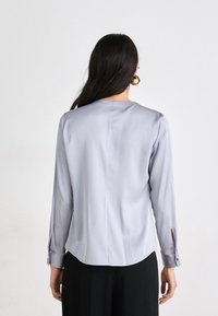 Vaaleanharmaa satiiniblouse pitkähihaisella, pyöristetyllä helman muotoilulla ja napillisilla hihoilla. Tasainen tekstuuri ja istuva leikkaus. Takaa otettu kuva.