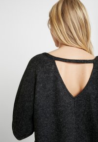 Pull en laine gris foncé avec un encolure large et une découpe en V à l'arrière. Présente un tissu texturé et doux, ainsi que des manches volumineuses.