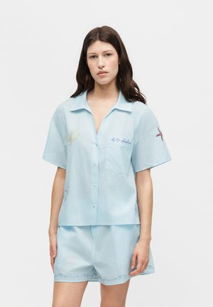 Γυναίκα που φοράει ανοιχτό μπλε κοντομάνικο πουκάμισο με κουμπιά και ταιριαστά σορτς με διακριτικά κεντημένα σχέδια εμπνευσμένα από τη θάλασσα.