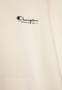 Crèmewitte sweatshirt van zachte stof, met een kangoeroezak en een zwart geborduurd "Champion"-logo op de voorkant.