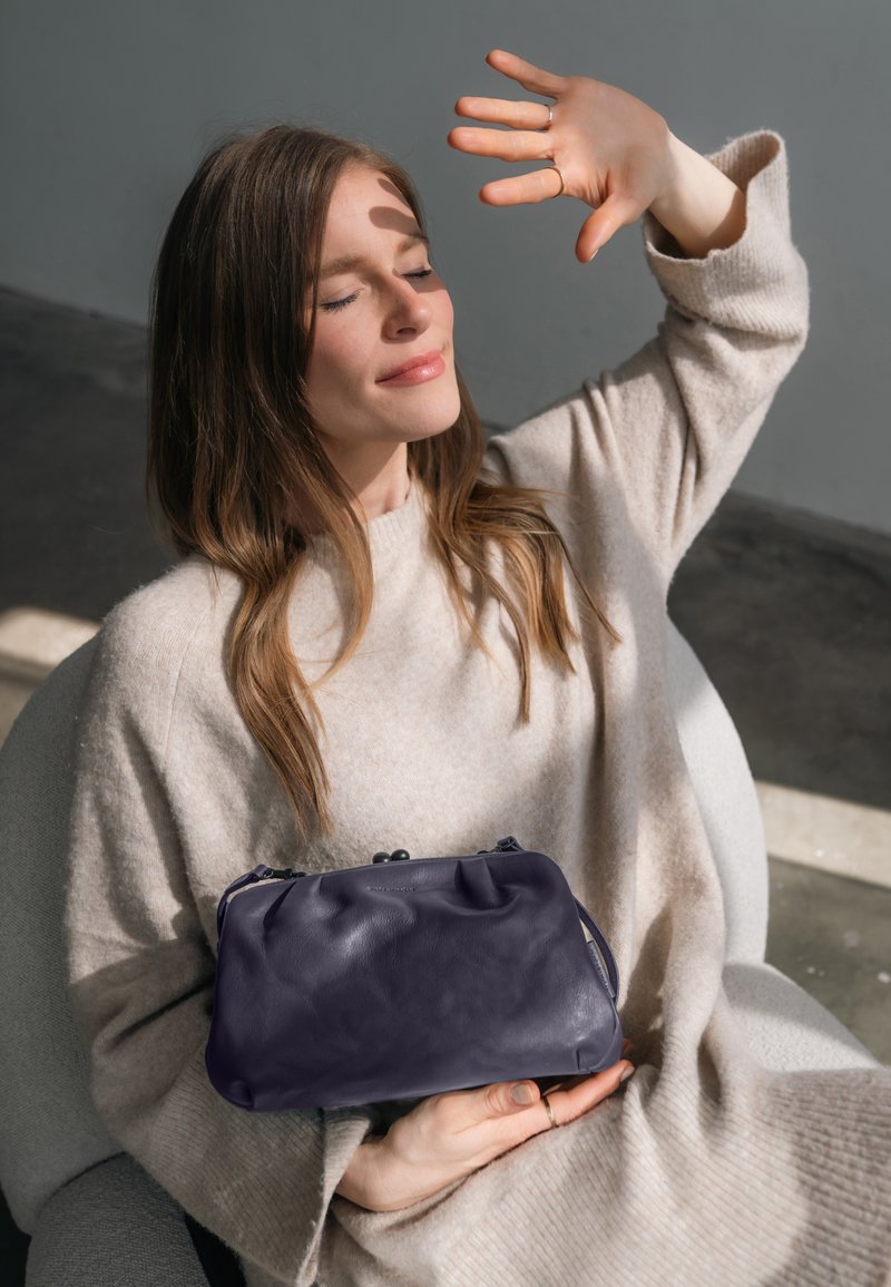 Lila Leder-Clutch mit runder Form, einem Deckel oben und einer glatten Textur, gehalten vor einem neutralen Pullover in einer sanft beleuchteten Umgebung.