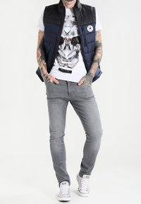 Gilet imbottito blu navy e nero con chiusura a zip, t-shirt bianca con grafica raffigurante un teschio, jeans grigi slim-fit e sneakers bianche.