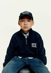 Chlapec sa priamo pozerá do kamery, má na sebe námornícky zipsový top s nápisom "EPIC FAIL" vytlačeným bielou farbou a čiapku NYC, sedí na čiernej stoličke.