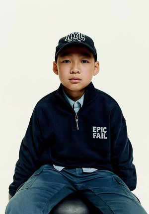 Ein Junge schaut direkt in die Kamera, trägt ein navyblaues Zip-Up-Shirt mit der Aufschrift "EPIC FAIL" in Weiß und eine NYC-Cap und sitzt auf einem schwarzen Hocker.