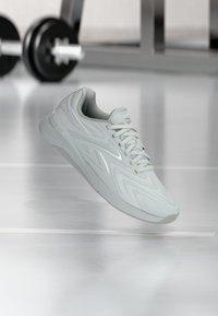 Αθλητικό παπούτσι Reebok σε ανοιχτό γκρι χρώμα με κορδόνια, αιωρούμενο πάνω από έναν καθρεφτισμένο δάπεδο γυμναστηρίου με θολά βάρη στο φόντο.