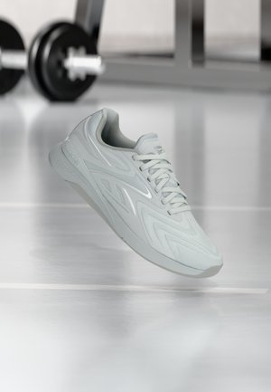 Lysegrå Reebok treningssko med lisser, svevende over et reflekterende treningsgulv med uklare vekter i bakgrunnen.