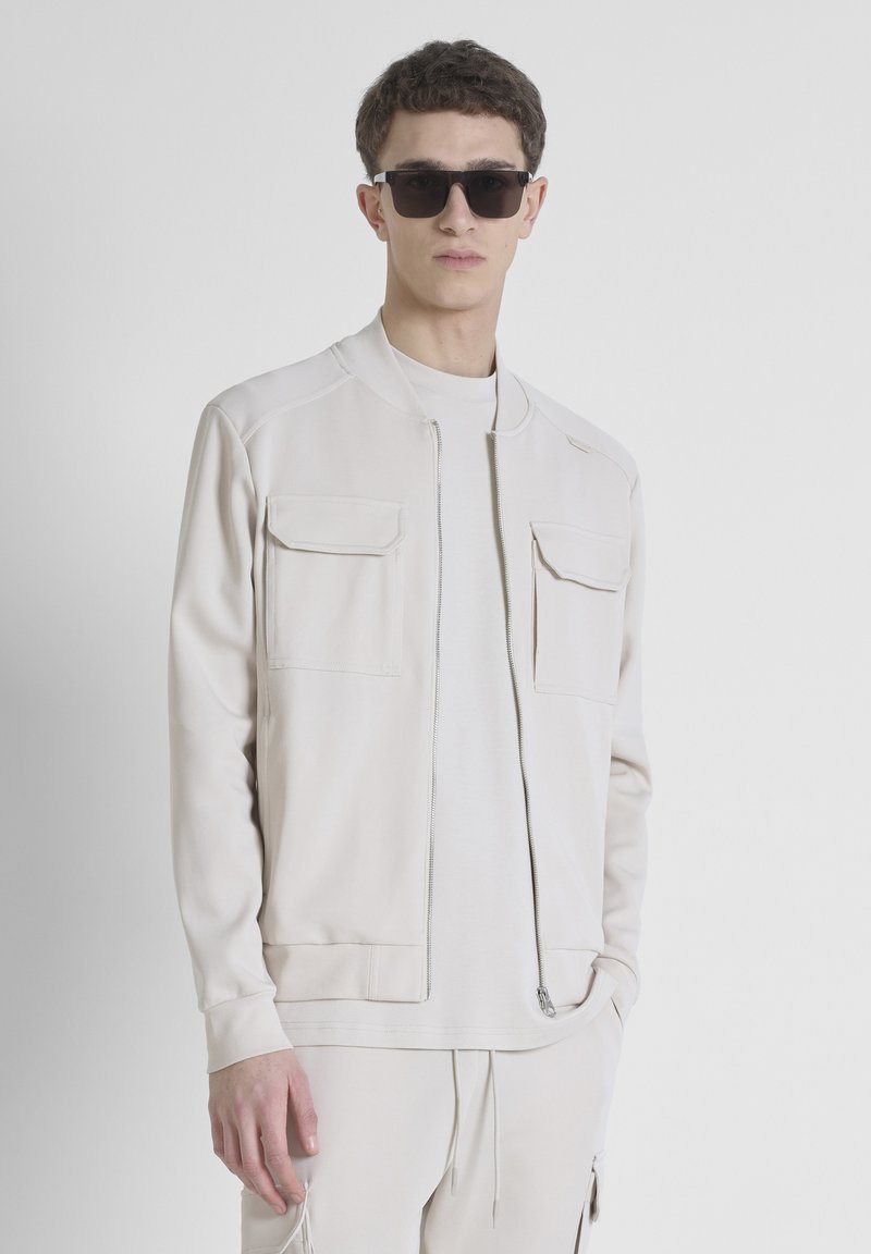 Chaqueta bomber beige con dos bolsillos frontales, cierre de cremallera, puños acanalados y cuello; combinada con una camiseta de color crema y pantalones a juego.