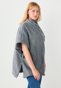 Poncho gris surdimensionné avec un col montant, une texture douce, des manches courtes larges et des fentes latérales pour faciliter le mouvement.