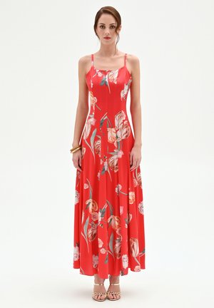 Femme debout de face, portant une robe longue fleurie rouge à fines bretelles et des sandales dorées, devant un fond blanc uni.