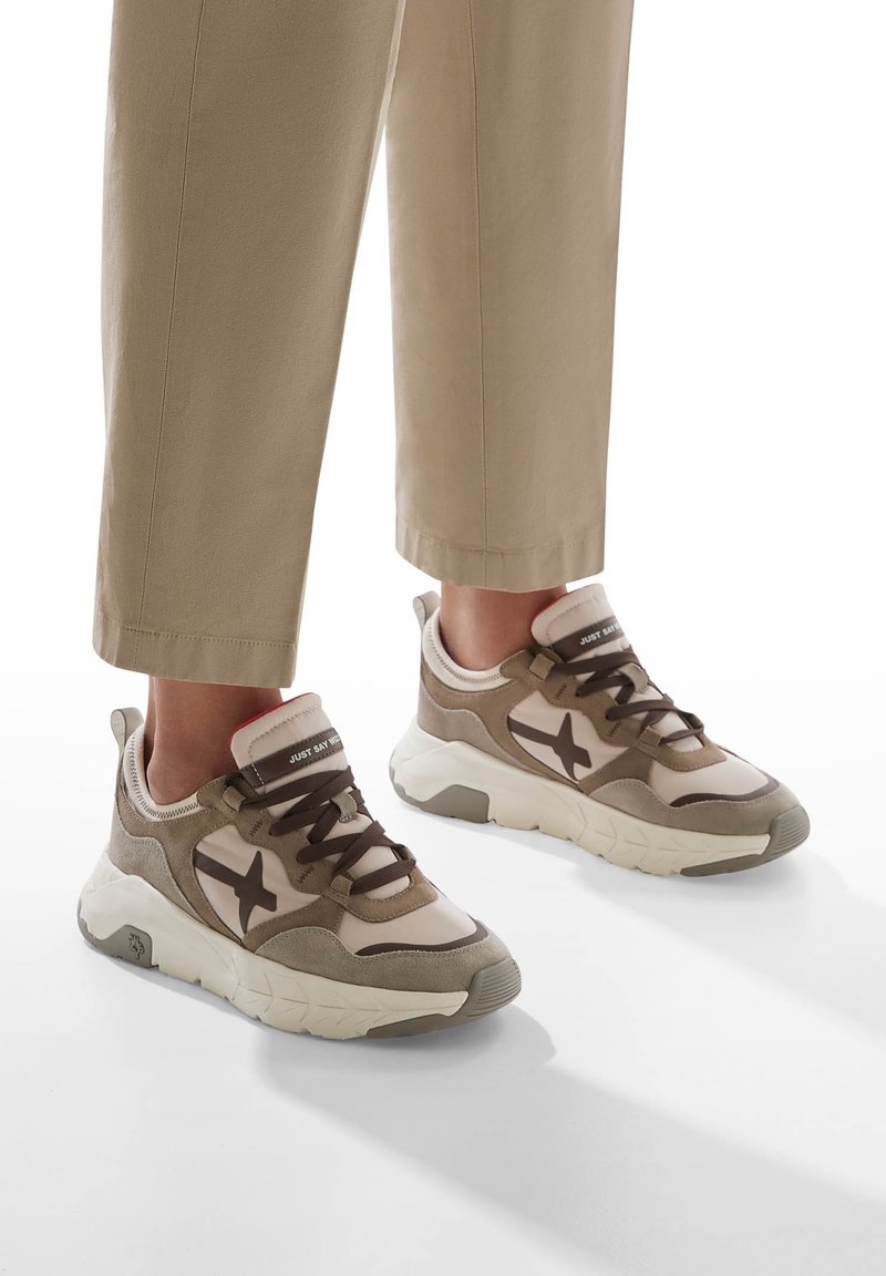 Pieds portant des baskets beige et marron décontractées avec des semelles blanches épaisses et des lacets foncés, associés à un pantalon beige court.