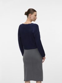 Kvinna med uppsatt hår i en knut, iklädd en marinblå stickad tröja och en grå knälång kjol, vänd något åt sidan mot en vit bakgrund.