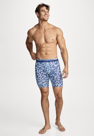 Muscular man standing barefoot wearing blue floral-patterned boxer briefs with "Jockey" waistband, smiling and looking to the side.