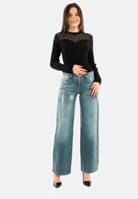 Top negro de manga larga con ribetes y secciones transparentes, combinado con vaqueros de pierna ancha azul claro, terminado con tacones altos negros brillantes.