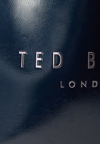 Ναυτικό μπλε δερμάτινο με ανάγλυφη ασημένια επιγραφή «TED BAKER LONDON.» Μινιμαλιστικός σχεδιασμός που εστιάζει στην ταυτότητα της μάρκας.