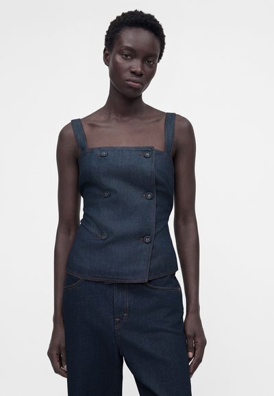 Νεαρή γυναίκα με κοντά μαλλιά που φοράει αμάνικη σκουρόχρωμη τζιν μπλούζα με κουμπιά και ταιριαστό σκουρόχρωμο τζιν παντελόνι σε απλό φόντο.