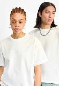 Dos personas llevan camisetas de color crema sueltas y ligeras. Una de las camisetas tiene un pequeño estampado cerca del escote. Las camisetas están hechas de una tela suave.