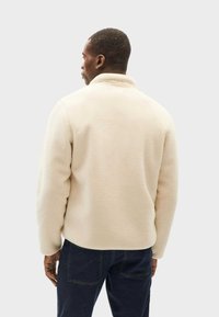 Fleecejacke in Off-White mit hohem Kragen und strukturierter Oberfläche. Sie hat einen entspannten Schnitt und lange Ärmel; passt gut zu dunklen Jeans.