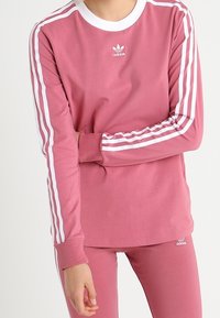 Rosa sweatshirt med vit krage och tre vita ränder på ärmarna, tillverkad av mjukt material, med en liten logotyp på bröstet.
