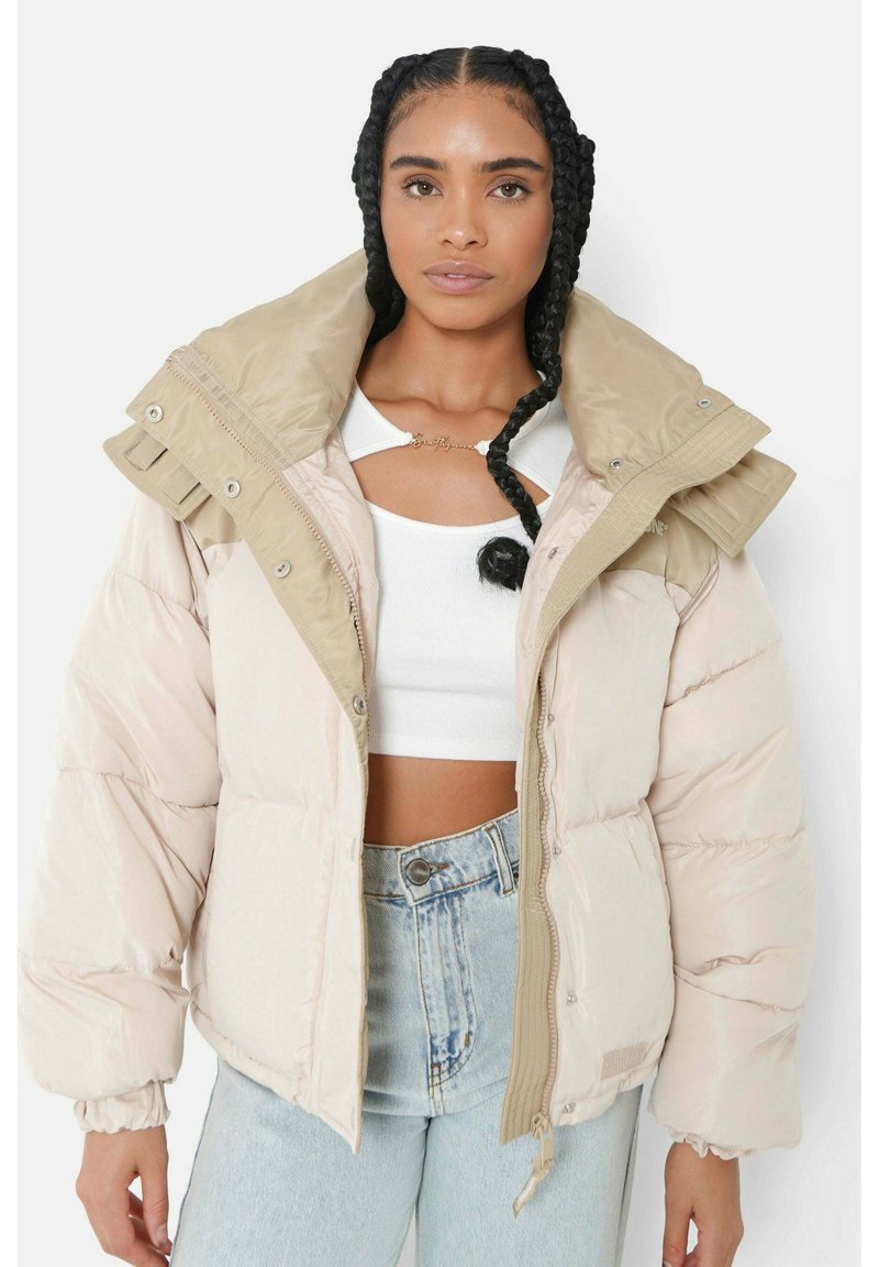 Giacca a vento beige con un grande colletto, dotata di bottoni a pressione, chiusura con zip e polsini a coste. Indossata sopra un top bianco.