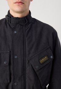 Casaco de algodão preto com gola alta, botões de pressão e um bolso no peito com um logótipo bordado a dourado. Tecido texturizado com costura vertical.