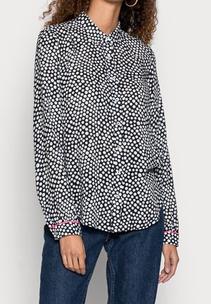 Mujer con blusa negra abotonada con lunares blancos y ribete rosa en los puños, combinada con vaqueros de mezclilla azul oscuro.