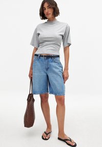 Pelēks īso piedurkņu tops kopā ar ziliem džinsu bermudu šortiem, melnu jostu un melniem čībām. Modelis tur rokās austu brūnu somu.