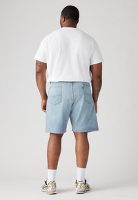 Homme portant un t-shirt blanc, un short en denim bleu clair, des chaussettes blanches et des baskets grises avec des accents dorés, se tenant devant un fond uni.