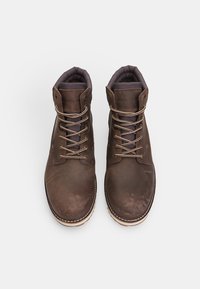 Botas de tobillo de cuero marrón con un acabado liso, punta redonda, forro textil oscuro y ojetes de metal para los cordones. Suela de goma con costuras en contraste.