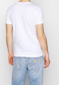 Camiseta de algodón blanca con mangas cortas, escote redondo, combinada con unos vaqueros de mezclilla azul claro que presentan una etiqueta de la marca en el bolsillo trasero.
