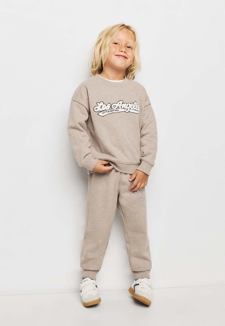 Enfant aux cheveux blonds portant un sweat-shirt beige et un pantalon de survêtement avec des baskets blanches, debout et souriant sur un fond blanc.