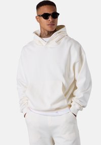 Hoodie blanc en tissu doux, avec une poche avant, une capuche à cordon de serrage et un branding minimal. Porté avec un bas assorti.