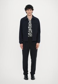 Črna jakna s ovratnikom, oblečena preko bele srajce, ki ima črn abstrakten vzorec. Črne hlače in čevlji dopolnjujejo videz.