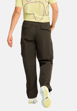 Persoon die wegloopt en donkere bruine cargobroek draagt, een lichtgele shirt met paarse strepen en witte sneakers met gele zolen.