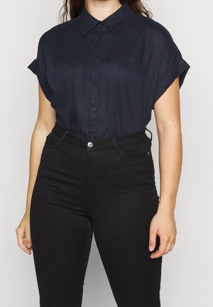 Femme portant une chemise à manches courtes bleu marine boutonnée, rentrée dans un pantalon noir taille haute, debout devant un fond uni.