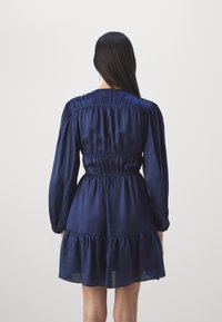 Námořnická modrá saténová šaty s dlouhými rukávy, nařasenými rameny a tiered volánkovým lemem. Obsahují elastický pás pro přiléhavý siluetu.