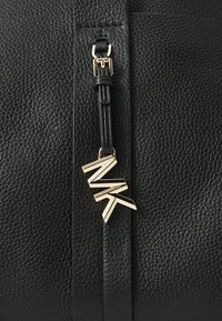 Sac en cuir noir texturé avec un tirette dorée et un pendentif en forme de logo géométrique. Le design comprend des coutures détaillées.