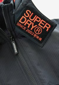 Giacca nera con tessuto a trama testurizzata a quadri. Include un audace patch ricamato con la scritta "SUPERDRY" e una chiusura frontale con zip.