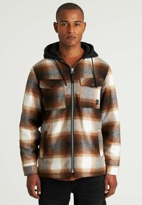 Chaqueta de franela con un patrón de cuadros en marrón y blanco, que cuenta con un cierre zip, dos bolsillos en el pecho y una capucha negra. Textura suave.