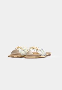 Sandales à glissière à motif floral en tissu léger avec une base beige. Elles présentent des accessoires en métal doré et une semelle plate en caoutchouc marron.