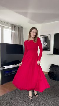 Rode jurk met lange mouwen, een vloeiende rok en een nauwsluitend lijfje, gemaakt van een zachte stof. Draagt zwarte sandalen met een gladde textuur.