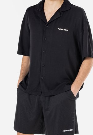 Mann trägt ein schwarzes kurzärmeliges Hemd mit Knöpfen und schwarze Netzshorts mit "PEGADOR" Logo auf beiden Kleidungsstücken, Hände in den Taschen.
