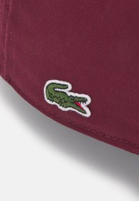 Raudonai rudas kepurė su tekstūruotu paviršiumi, su žaliu krokodilo logotipu, siuvinėtu raudonomis detalėmis. Tinka kasdieniam nešiojimui.