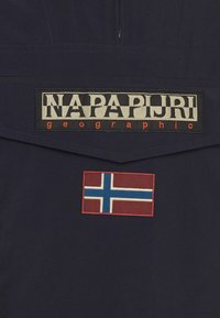 Szczegół kurtki Napa Pijri, z granatowego materiału, dużym blokowym logo oraz haftowaną łatą z norweską flagą.