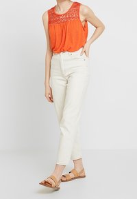 Pomarańczowy bezrękawnik z koronkowym wykończeniem, połączony z białymi dżinsami o wysokim stanie i brązowymi sandałami. Prosty, luźny fason i swobodny wygląd.
