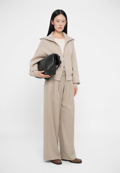 Beige ribbet cardigan med bred krage, kombinert med beige plissert bukser. Svart lærveske holdt under armen; brune sko fullfører antrekket.