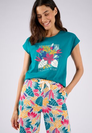 T-shirt a maniche corte color turchese con una grafica floreale e il testo bianco "NOTTE TROPICALE", abbinato a pantaloni multicolori con una fantasia di foglie e fiori.