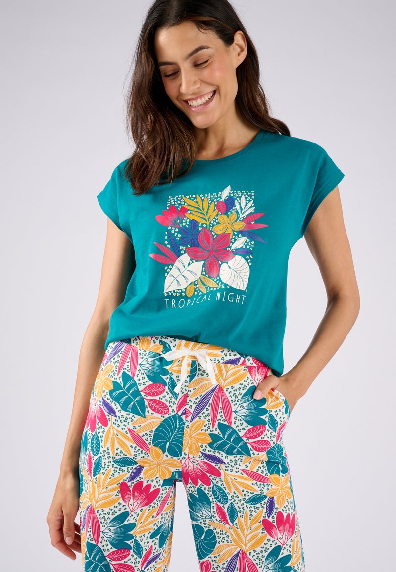 T-shirt a maniche corte color turchese con una grafica floreale e il testo bianco "NOTTE TROPICALE", abbinato a pantaloni multicolori con una fantasia di foglie e fiori.