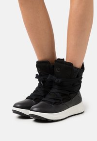 Bottines mi-hauteur noires et grises avec un dessus en cuir texturé et en daim, présentant un col rembourré, de lacets épais et une semelle en caoutchouc blanc.