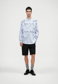 Camisa de botão riscada em azul claro com múltiplos laços decorativos, combinada com calções pretos. Modelo a usar sapatos pretos e meias cinzas.